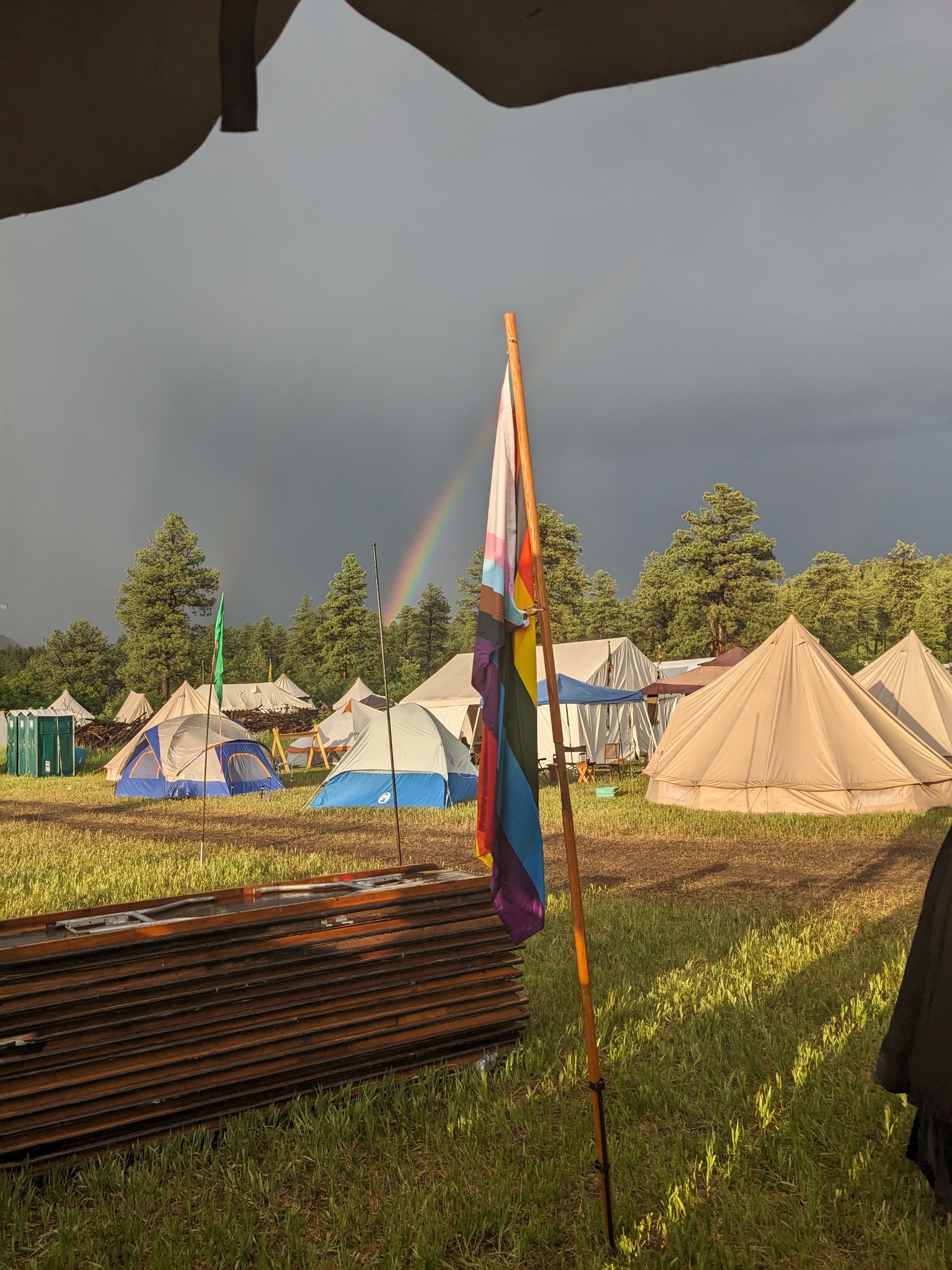 Picture of a pride flag in front of tents. There are clouds in the sky and a rainbow.